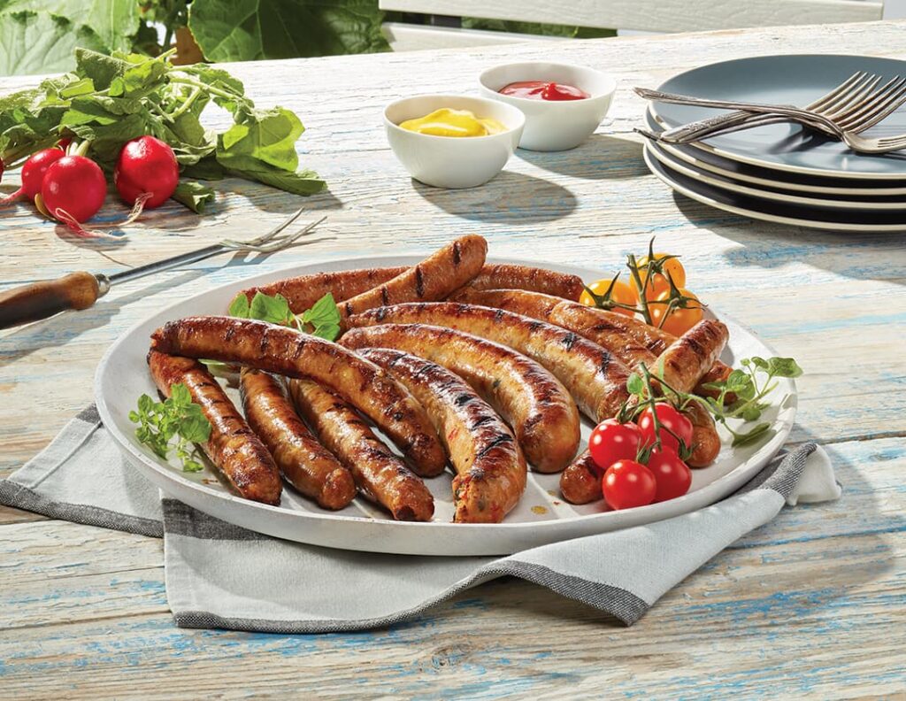 Plusieurs saucisses Ferme des Voltigeurs dans une assiette sur une table extérieure accompagnées de tomates et d'herbes.