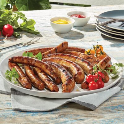 Plusieurs saucisses Ferme des Voltigeurs dans une assiette sur une table extérieure accompagnées de tomates et d'herbes.