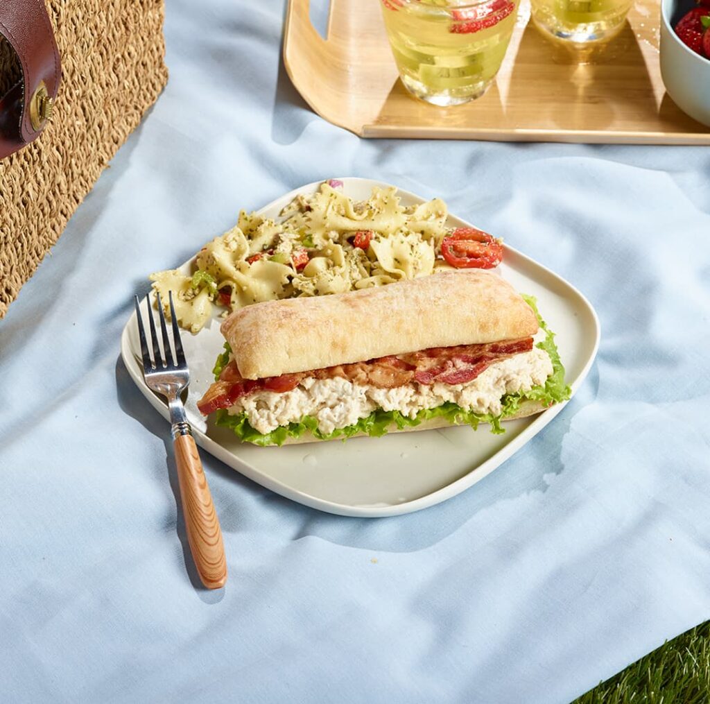 Caesar Sandwich with Ferme des Voltigeurs Grain-Fed Chicken Spread, served with a pasta salad in a picnic setting.