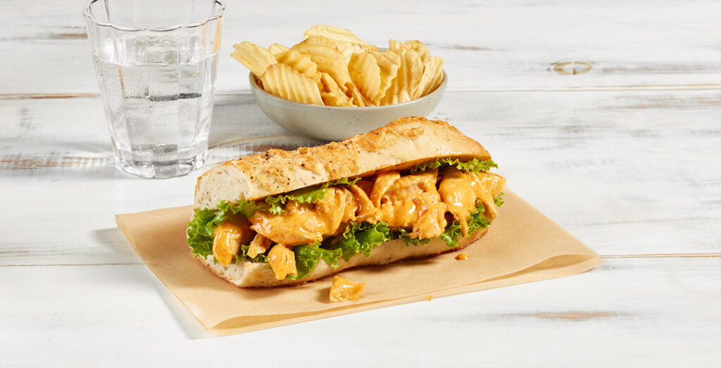 Sandwich au poulet chipotle de Ferme des Voltigeurs, photo ambiance sur table grise et accompagnement de chips. / Ferme des Voltigeurs Chipotle Chicken Sandwich, lifestyle photo on a gray table with a side of potato chips.