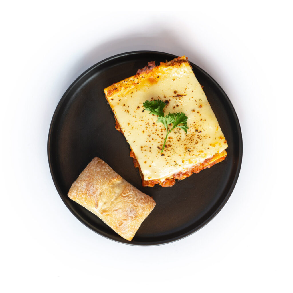 Lasagne au poulet de Ferme des Voltigeurs sur fond blanc, accompagné d'un pain ciabatta. / Ferme des Voltigeurs Chicken Lasagna on a white background, served with a ciabatta bun.